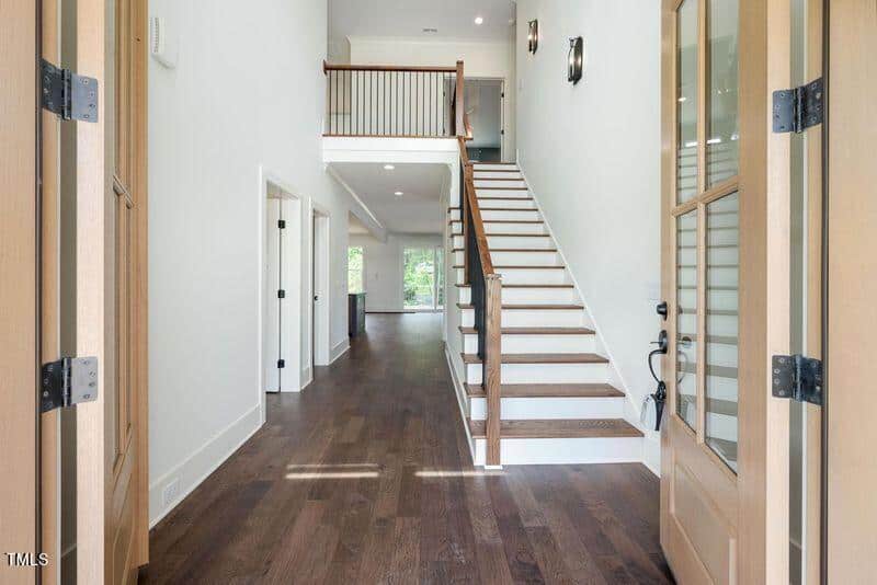Spacious interior of a modern home with wooden floors and a staircase leading to an upper level. Glass doors open into the bright hallway, and natural light floods the space from windows and a balcony railing above.