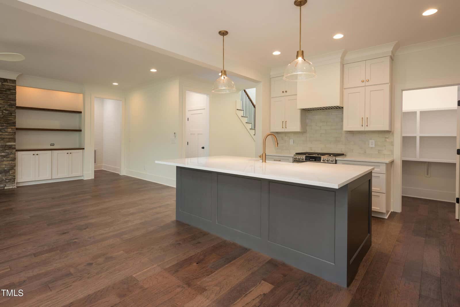 A modern kitchen with a large island featuring a white countertop and gray base. The space includes wooden floors, pendant lighting, a gas stove, and light-colored cabinets. An adjacent room has built-in shelves and stone accents on a wall.