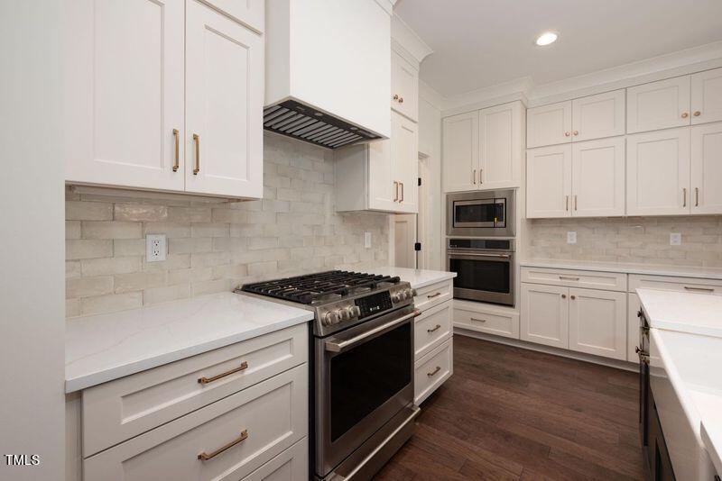 Modern kitchen with white cabinets, light beige tiled backsplash, dark wood flooring, and stainless steel appliances, including a stove and double ovens. Quartz countertops and recessed lighting enhance the space.