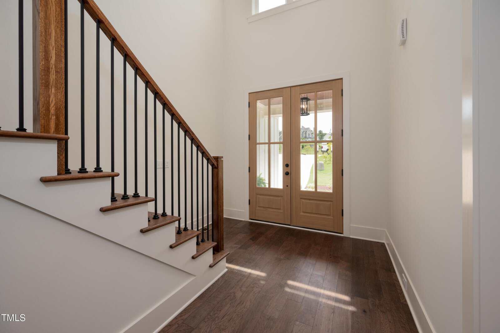 A bright, modern entryway featuring a wooden staircase with black railings on the left. Double wooden front doors with glass panels let in natural light, illuminating the dark hardwood flooring. White walls add to the airy atmosphere.