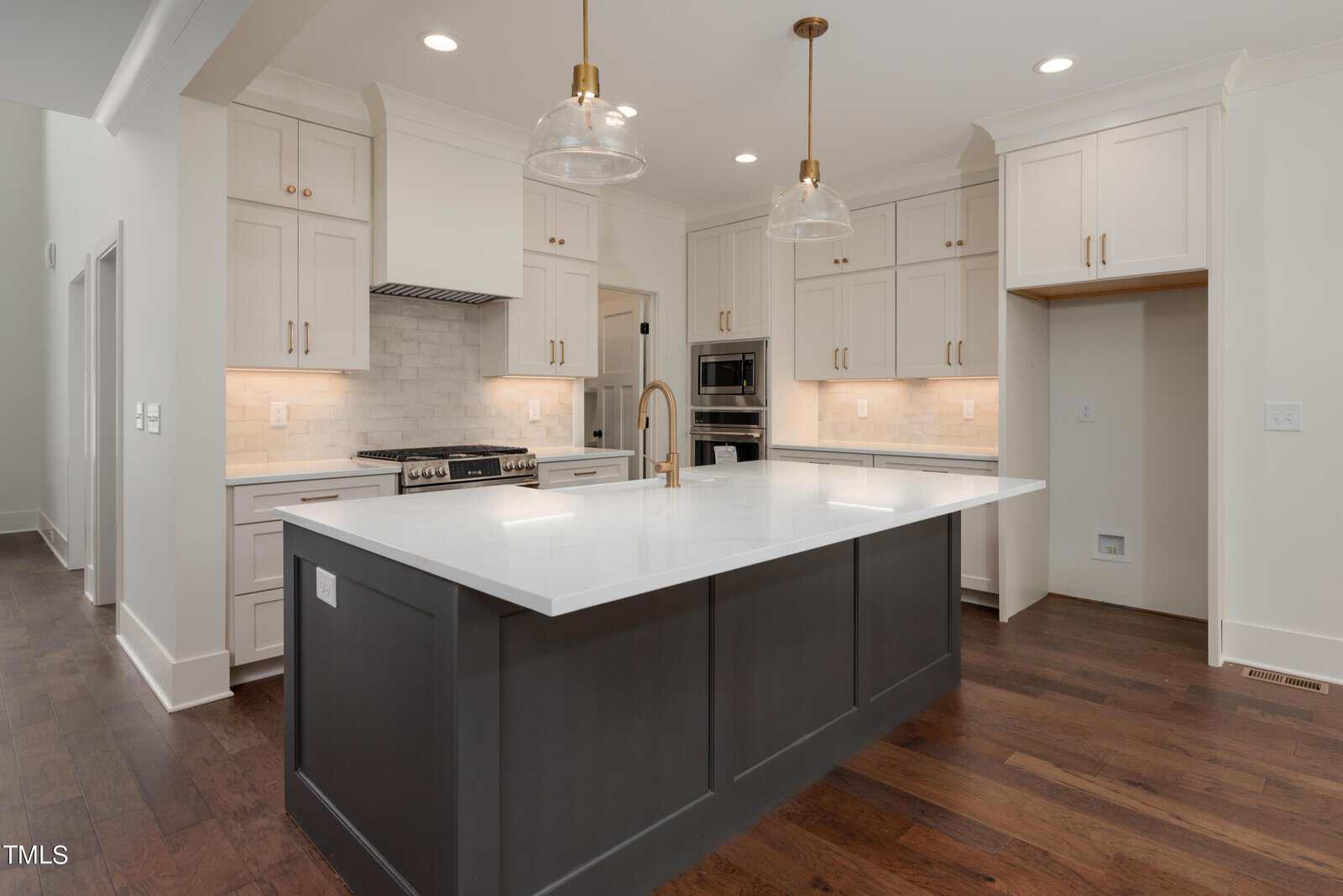A modern kitchen featuring a large island with a white countertop and dark base. The background includes white cabinets, a stainless steel oven, a gas stove, and two pendant lights hanging over the island. Dark hardwood floors complete the look.