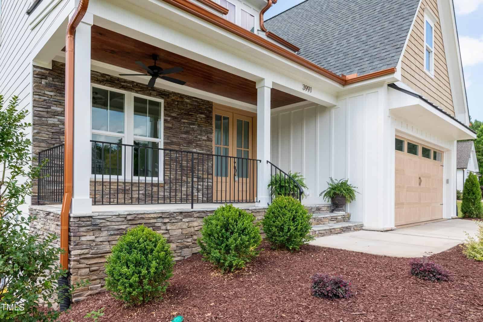 A house with a stone and white siding exterior, featuring a front porch with black railing and a wooden door. There are shrubs in the landscaped front yard and a ceiling fan on the porch. A garage is visible on the right.