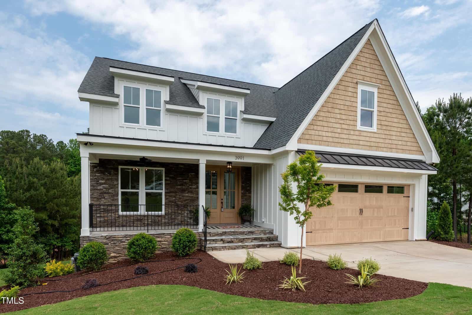 A modern two-story house with a mix of stone and wood siding. It features a steep roof, a front porch with black railing, and a double garage. The landscaped yard includes grass and small shrubs, set against a backdrop of green trees and a cloudy sky.