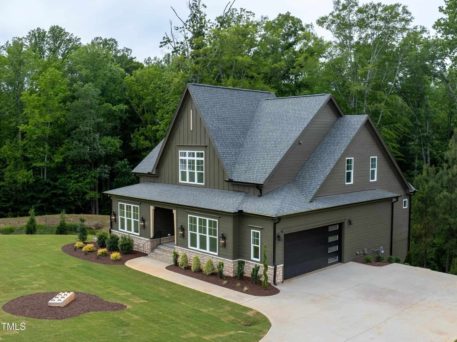 A modern two-story house with a dark gray exterior, stone accents, and a spacious driveway. The yard is landscaped with shrubs and a small garden. Surrounded by lush green trees under a cloudy sky.