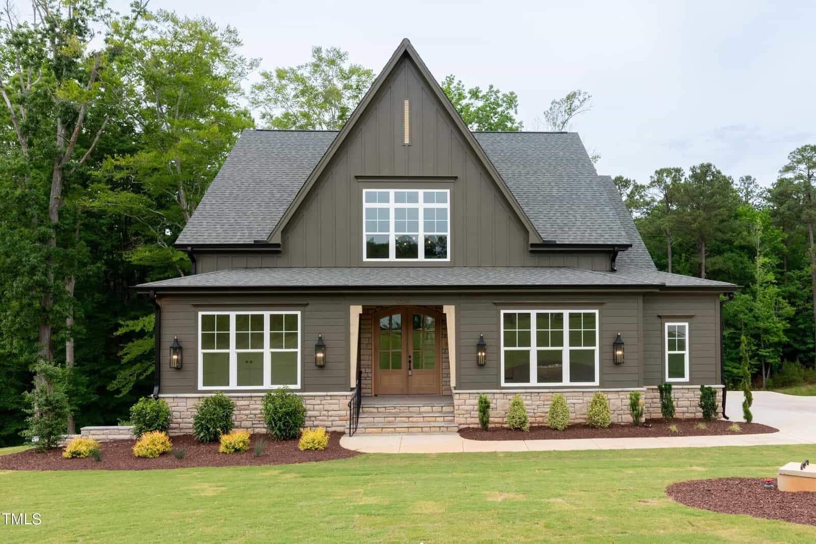 A modern two-story house with a steep, gabled roof, large windows, and a covered entrance. The exterior is painted dark gray with stone accents. The house is surrounded by a well-maintained lawn and lush greenery.