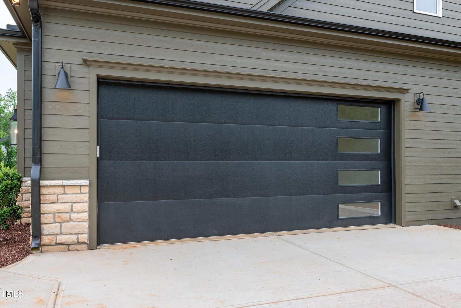 A modern garage door with horizontal panels, featuring three small rectangular windows on the right side. The exterior wall is a soft green with brick accents. Two outdoor lights are mounted on either side of the door.
