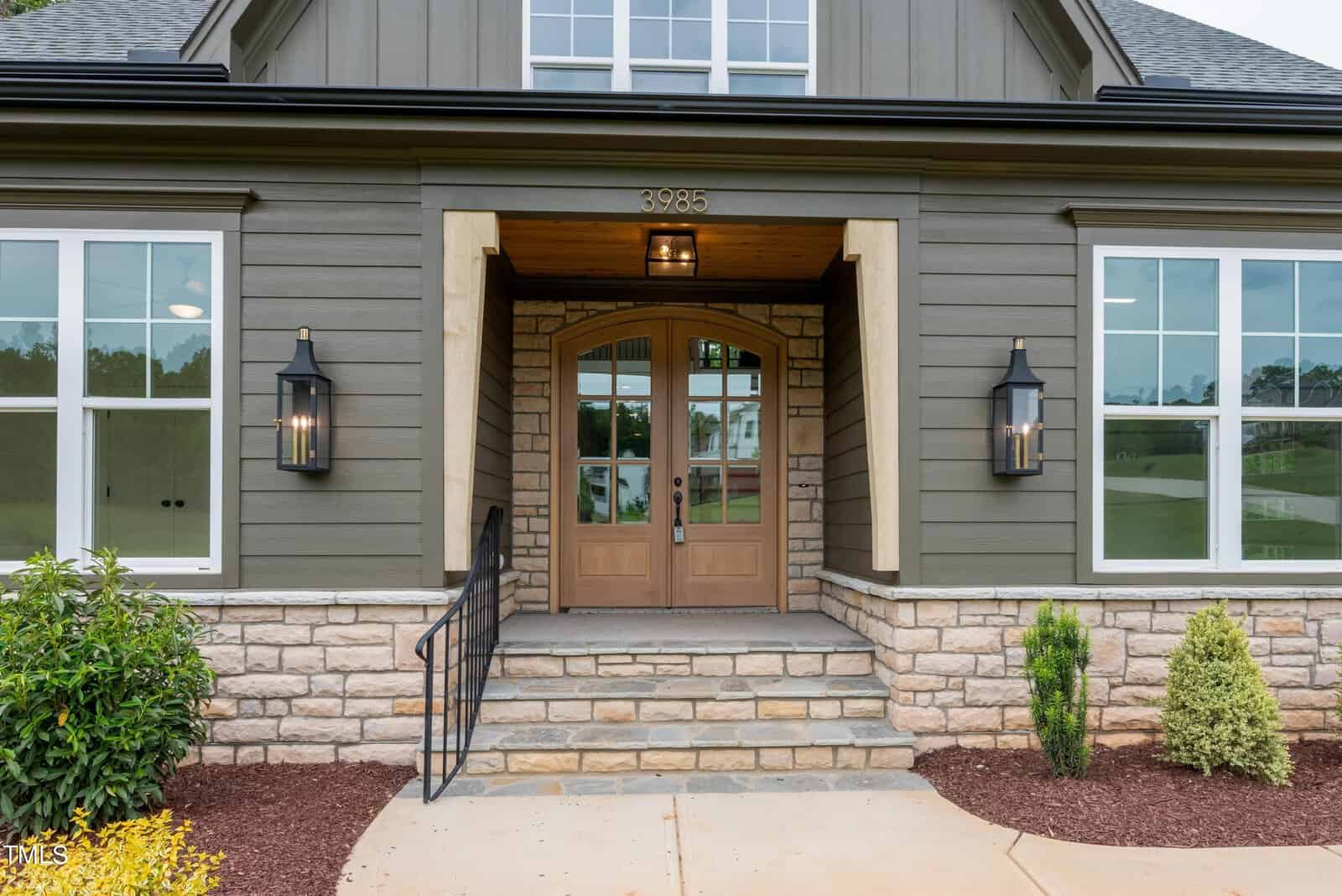 A modern house entrance featuring wooden double doors flanked by large lanterns. The exterior includes gray siding, stone accents, and multiple windows. The steps leading to the door are bordered by black railings, and there's landscaped greenery.
