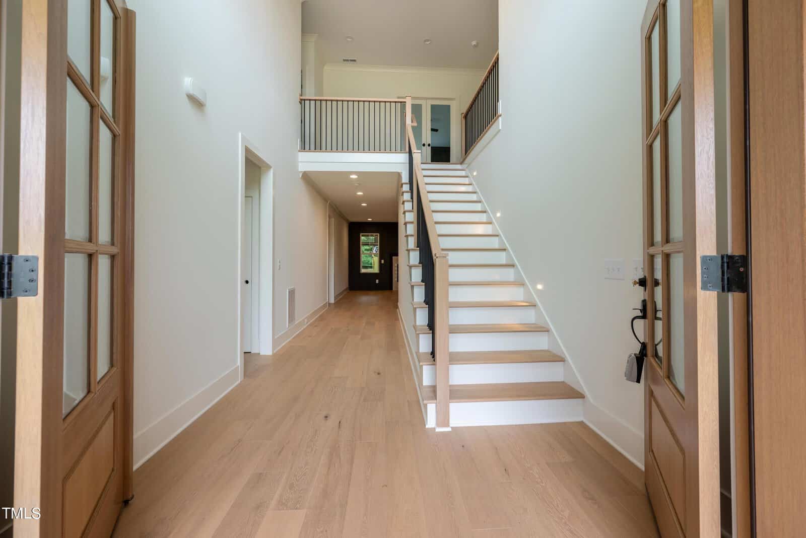 A spacious entrance hallway with light wooden flooring, double wooden doors, and a staircase with white risers and wooden treads leading to an upper floor. The walls are white, and a railing overlooks the entrance from above.