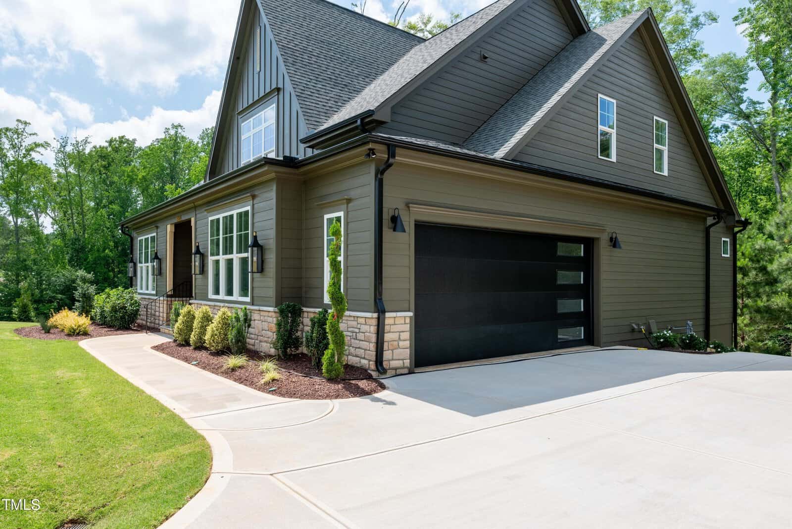 A modern two-story house with gray siding, large windows, and a black garage door. The driveway is wide and clean, and the garden is landscaped with small bushes. Trees and a clear sky are in the background.