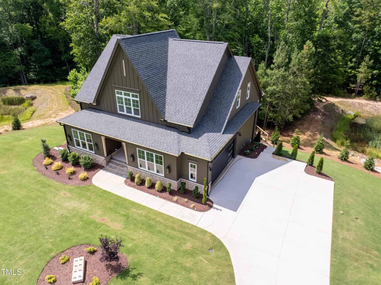 Aerial view of a large, modern house with dark gray roofing and a green exterior. It features a manicured lawn, a spacious driveway, and landscaped garden beds. Surrounded by lush trees, the home is set in a serene, green environment.
