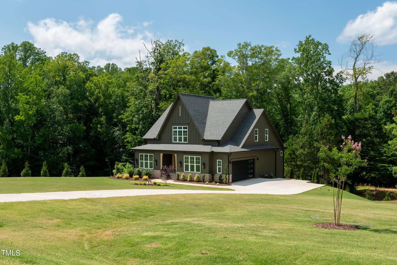 A two-story dark gray house with a steep roof, large windows, and a spacious driveway is surrounded by lush green trees and a manicured lawn under a partly cloudy sky.