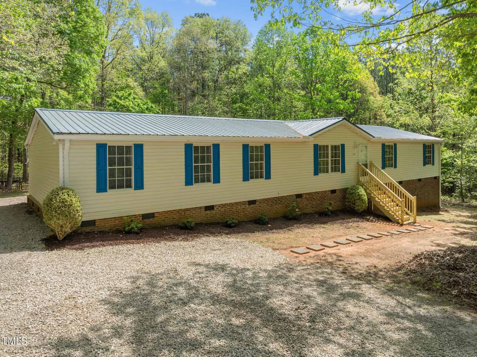 A beige mobile home with blue shutters and a metal roof sits peacefully at 3196 Loblolly Lane, Wake Forest, surrounded by lush green trees. A gravel driveway leads to the front steps, where a small path outlined with stones enhances the serene, wooded setting.