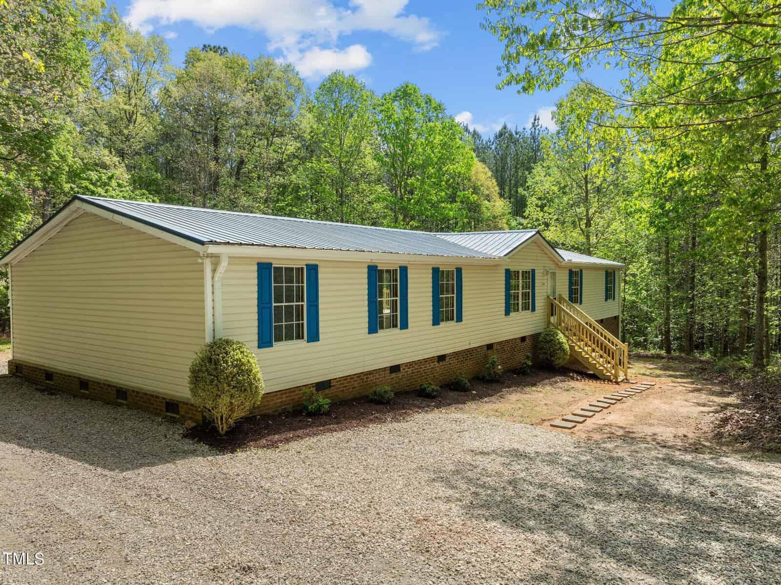 A beige manufactured home with blue shutters and a metal roof sits in a wooded area under a blue sky with clouds. Wooden steps lead to the entrance from a gravel path, and small bushes line the front of the house.