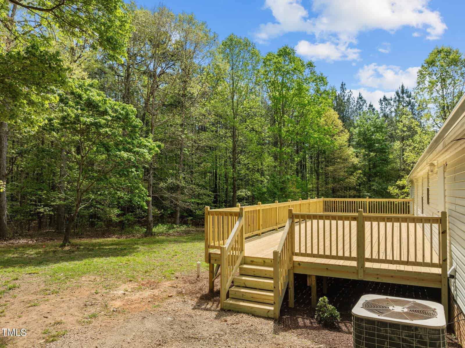 A sunny backyard features a spacious wooden deck with railings, leading down to a grassy area. The scene is surrounded by lush green trees under a blue sky with scattered clouds. An HVAC unit is visible next to the house.