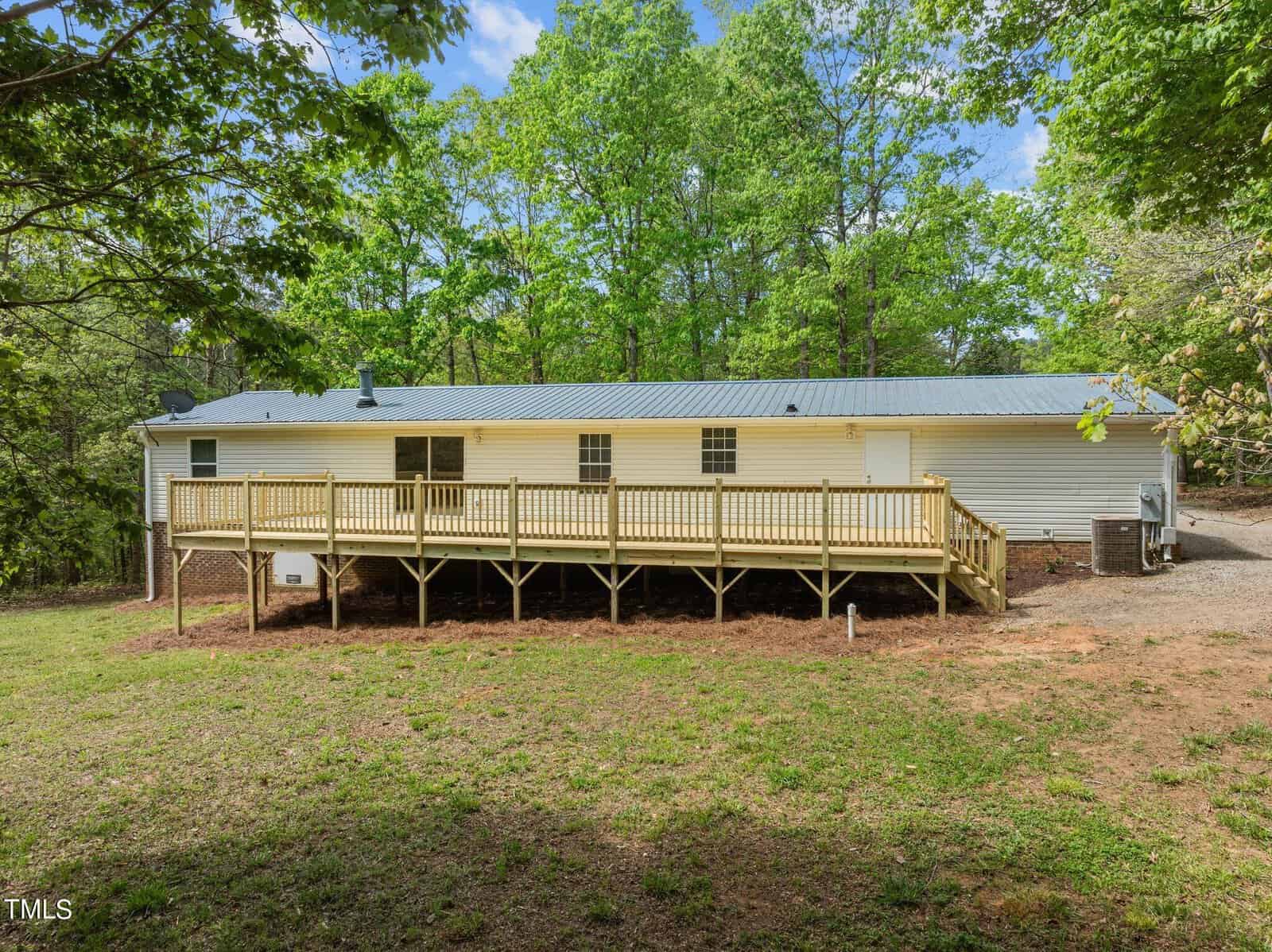 A single-story house with a metal roof and a spacious wooden deck, surrounded by a lush green forest. The yard is grassy with some bare patches, and the sky is clear and blue overhead.