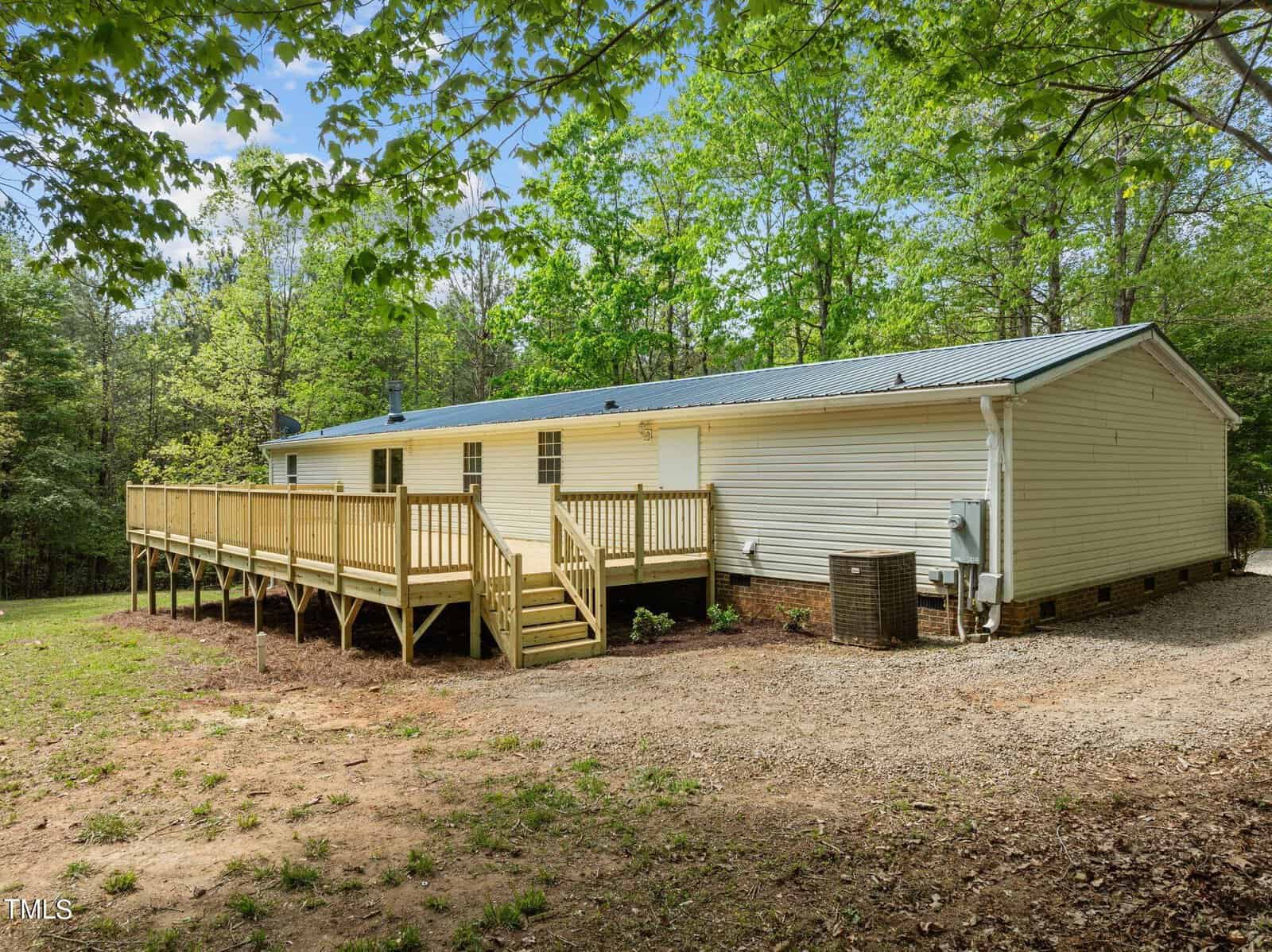 A single-story, light-colored house with a metal roof is surrounded by trees. It features a long wooden deck and ramp on the side and a gravel area in the foreground. The setting is outdoors with lush greenery and a partly cloudy sky.