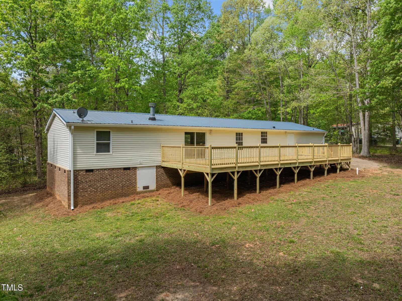 A single-story white house with a metal roof is surrounded by trees. It features a wooden deck along the front, and the foundation is made of red brick. The yard is spacious with grass and some mulch. The sky is clear and blue.