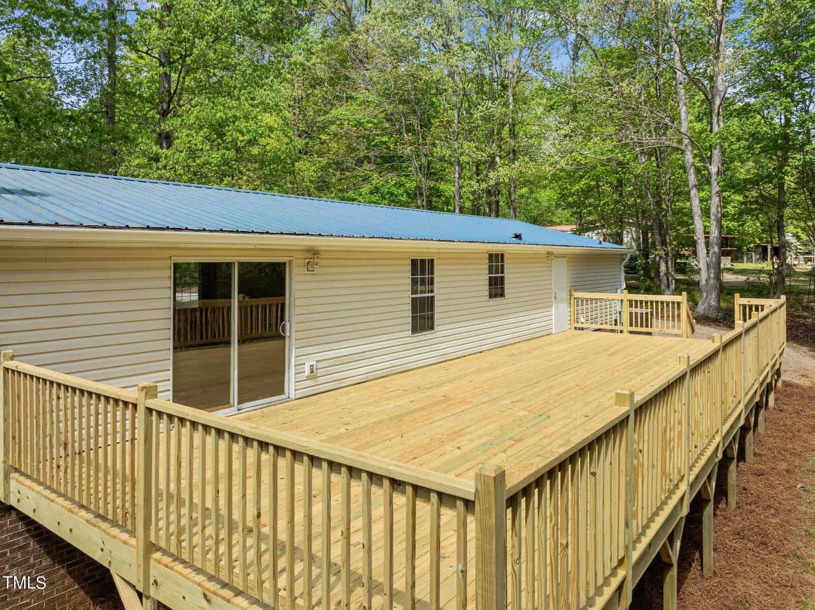 A newly built wooden deck attached to a single-story home with a metal roof. The deck features railings, and the house has sliding glass doors. Surrounding the area are lush green trees and a wooded landscape.