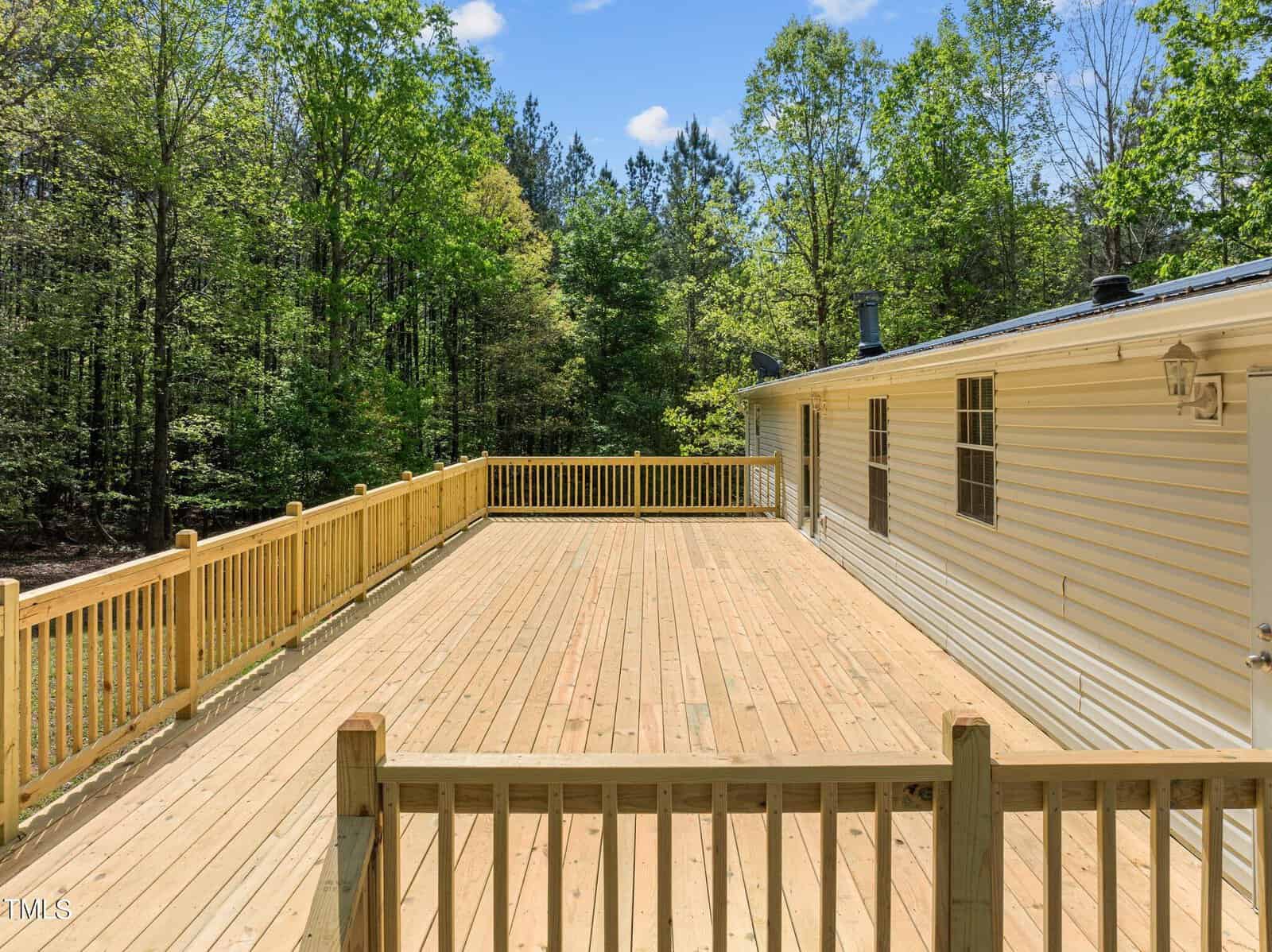 A spacious wooden deck is attached to a white mobile home. The deck is enclosed by a simple wooden railing. Surrounding the area are lush green trees, under a bright blue sky with a few clouds.