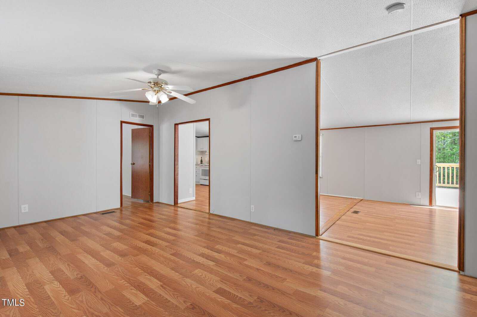 Spacious, unfurnished room with wood flooring and light gray walls. Features a ceiling fan, open doorway to a kitchen, and a door leading to another room. Large window provides natural light, overlooking a green outdoor area.