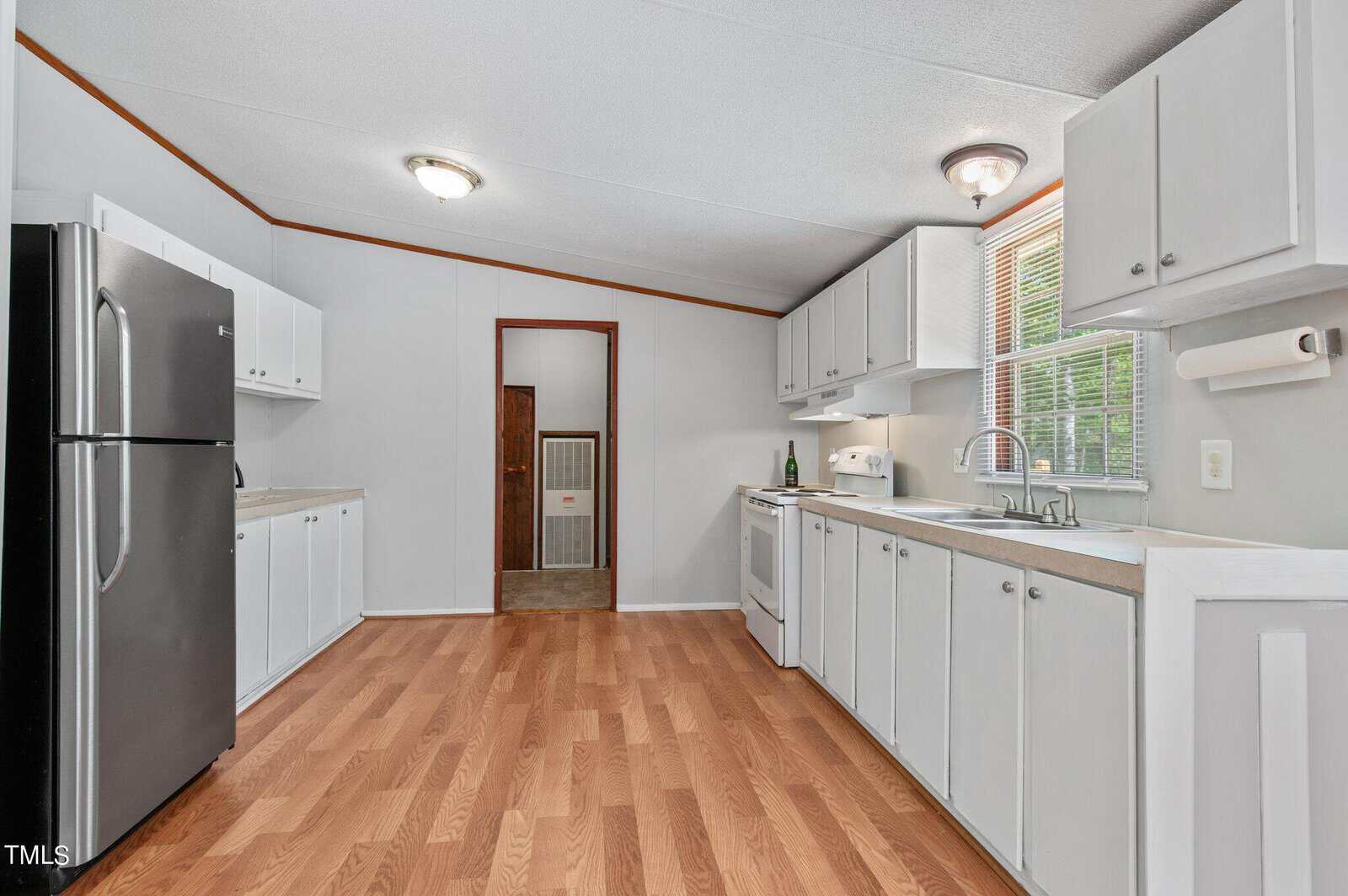 A bright, modern kitchen with white cabinets, a stainless steel refrigerator, and a white stove. Wooden floors and a large window provide warmth and natural light. Doorway leads to another room in the background.