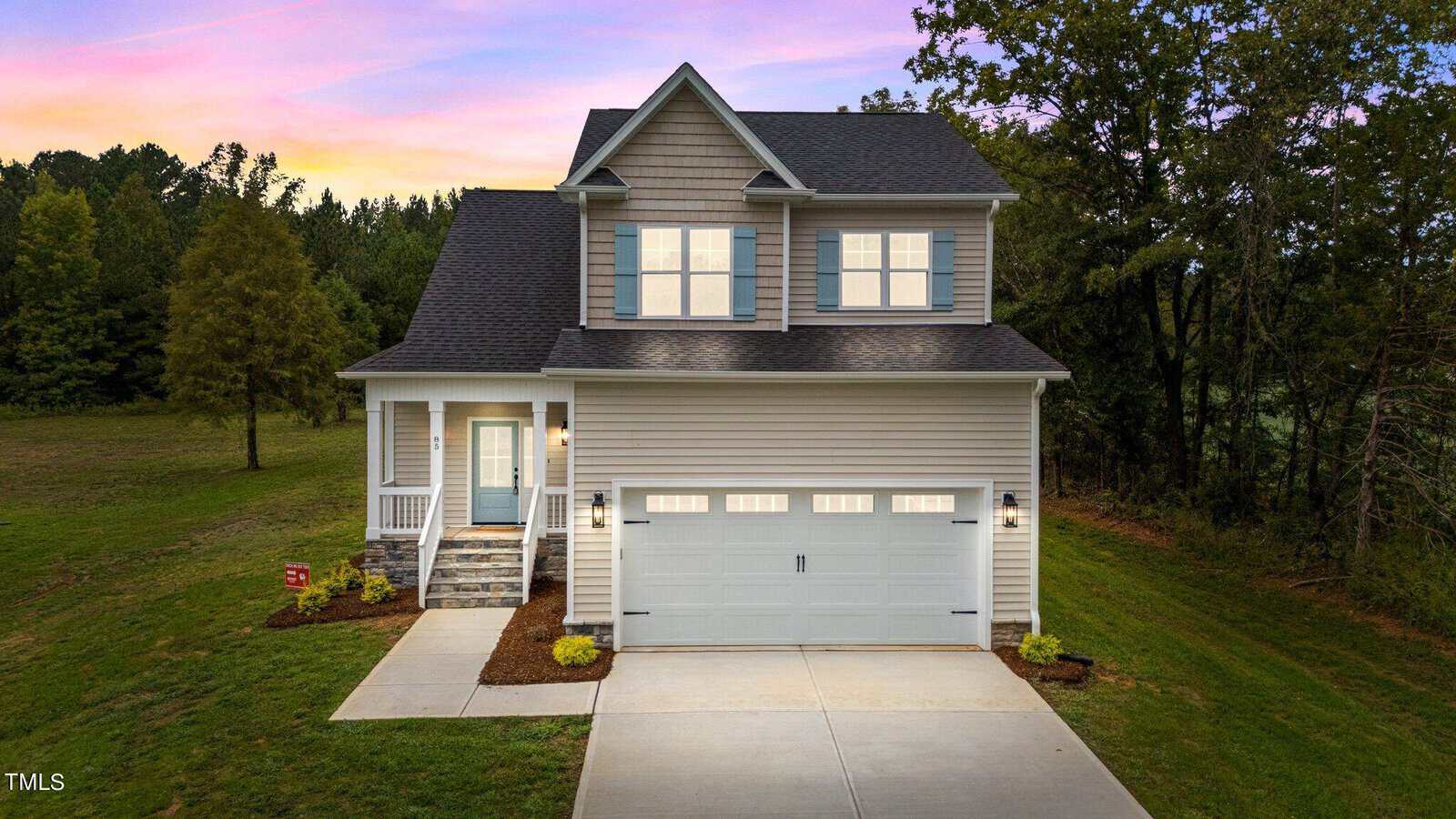 A two-story modern house with a gray exterior, white trim, front porch, double garage, and a well-kept lawn, set against a backdrop of trees and a colorful sunset sky.