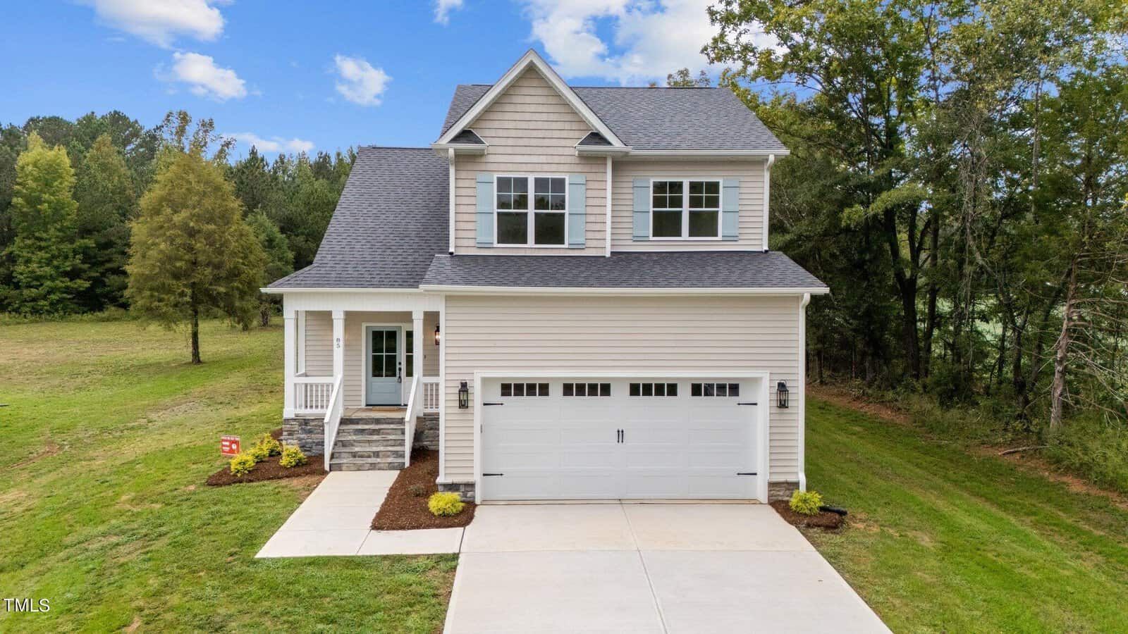 A two-story beige house with a gray roof, white trim, and attached two-car garage. The house has a small front porch with stairs, sits on a grassy lot, and is surrounded by trees under a blue sky with clouds.