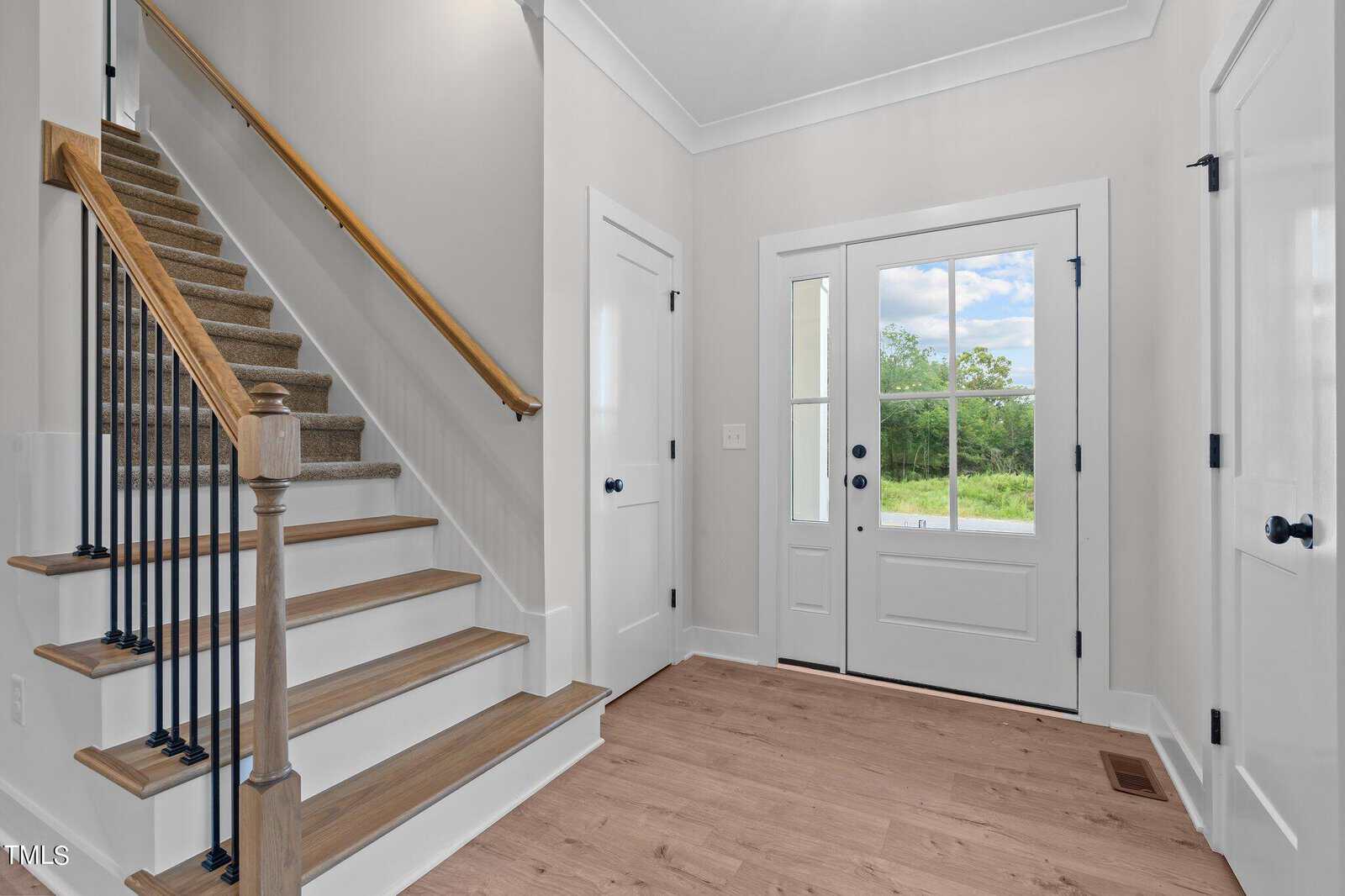 A bright entryway with wooden stairs and black railings on the left, white double doors with glass panes opening to a green view, white walls, and light wood flooring. Two closed white doors are on the right.