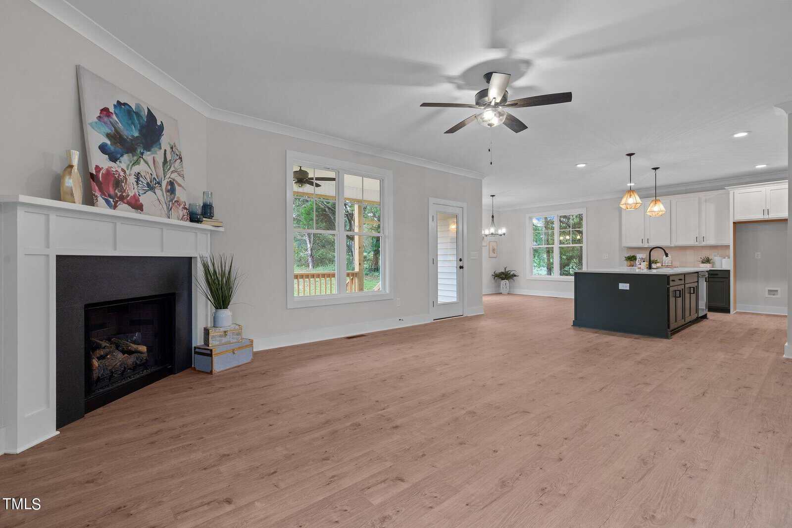 Open-concept living space with light wood floors, a ceiling fan, white walls, a fireplace with art above, large windows, and a modern kitchen with an island and pendant lights in the background.