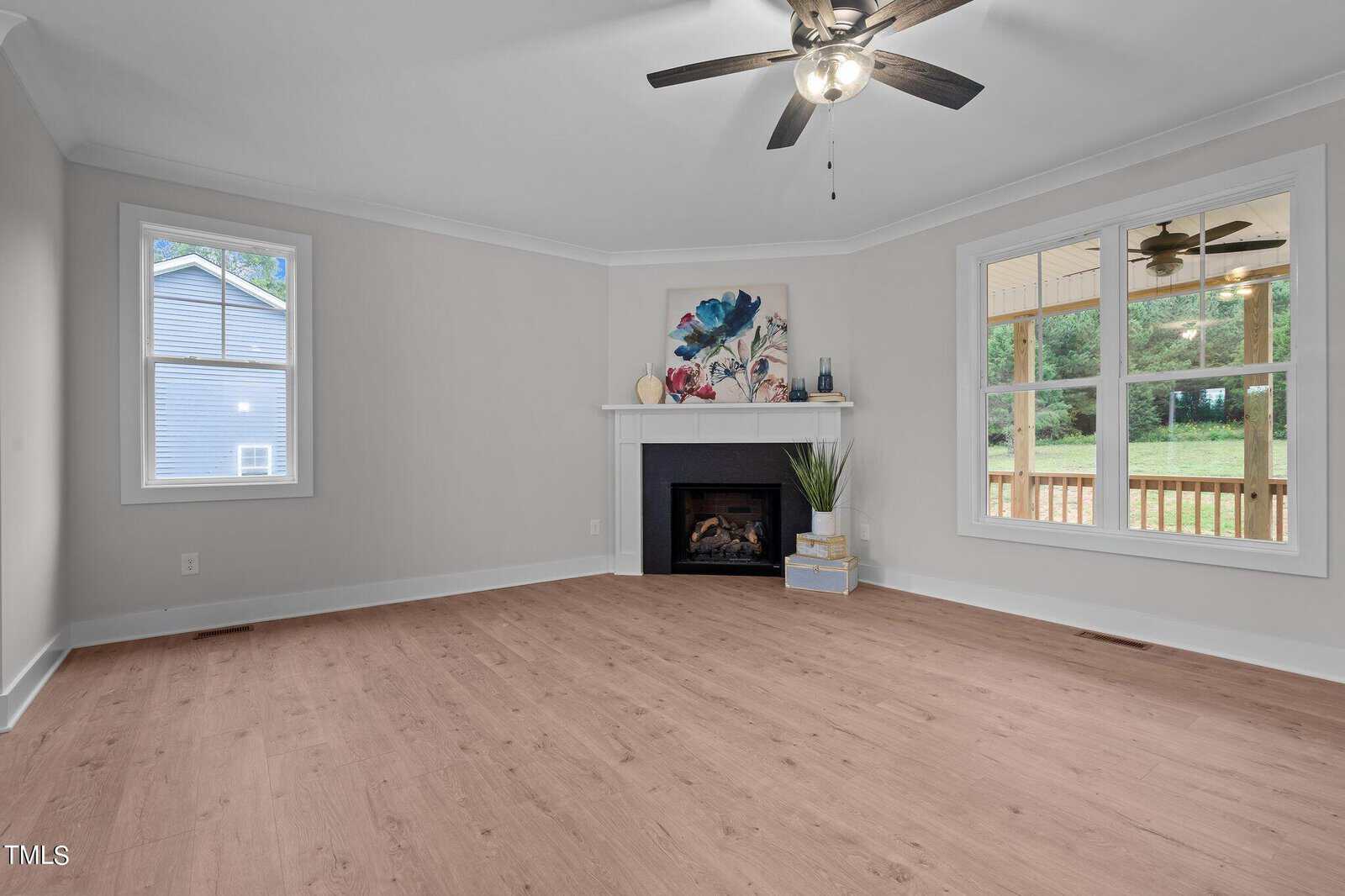 A bright, empty living room with light wood floors, a ceiling fan, a fireplace with decor, and large windows showing a porch and greenery outside.