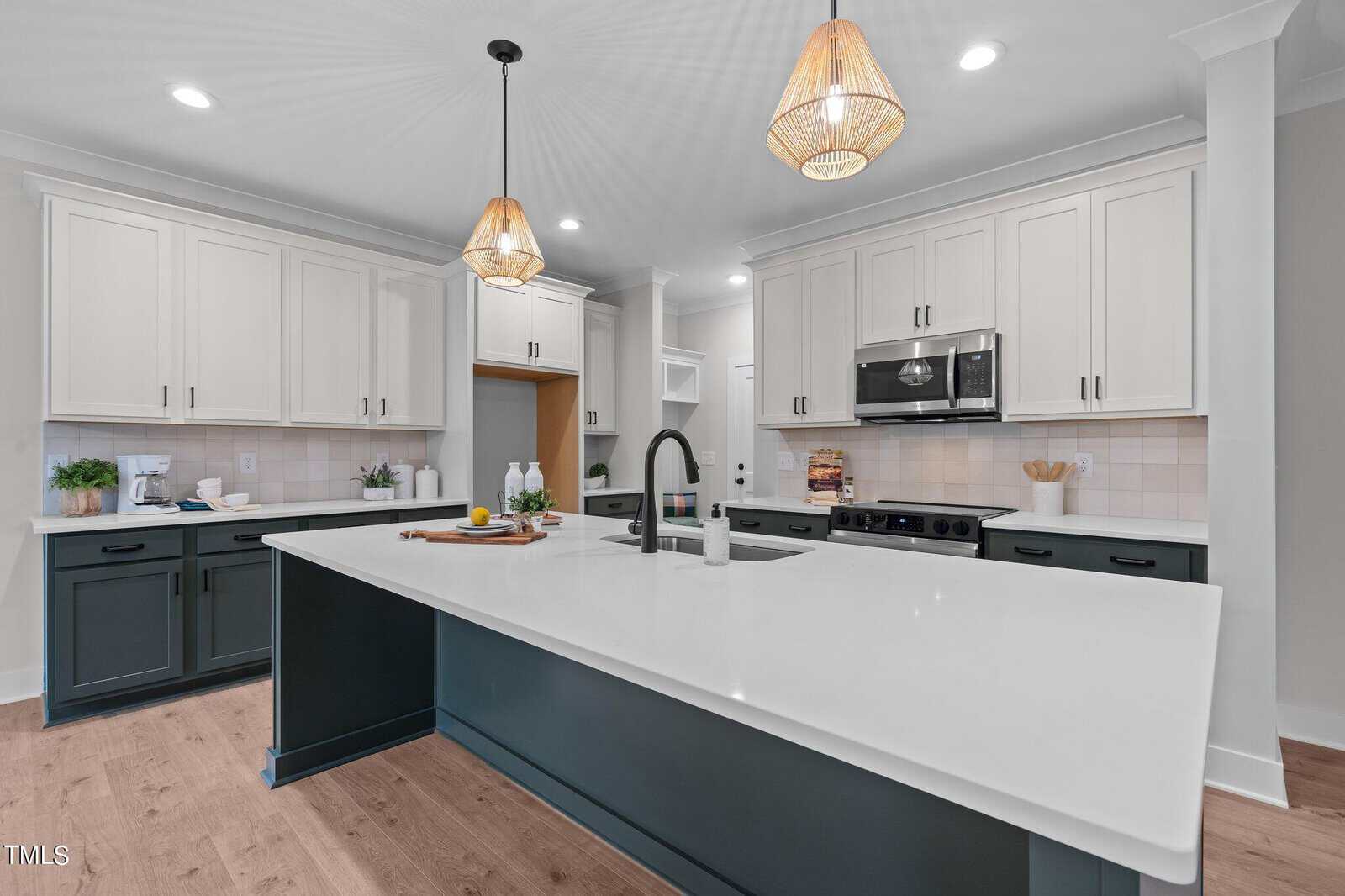 Modern kitchen with a large white island, dark lower cabinets, white upper cabinets, stainless steel appliances, two pendant lights, and light wood flooring. The space is clean and well-lit.