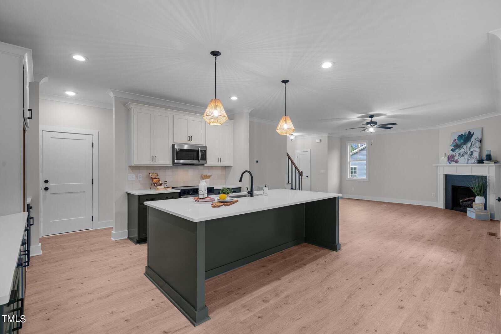Modern open-concept kitchen and living area with light wood floors, white walls, a large dark green island, stainless steel appliances, pendant lights, and a cozy fireplace with a painting above it.