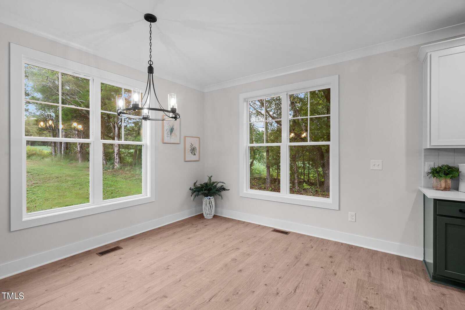 Bright dining area with large windows showing green outdoor scenery, light wood floor, white walls, a black chandelier, wall art, and a potted plant in the corner next to a modern kitchen counter.
