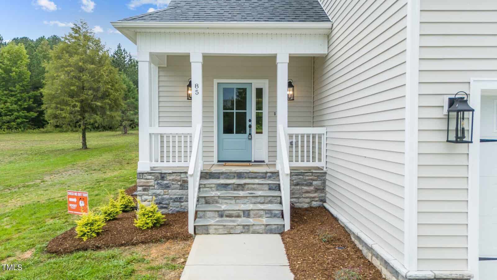 A small front porch with white railings, stone steps, and a light blue door on a beige house. There are shrubs and mulch along the walkway, and a grassy lawn with trees in the background.