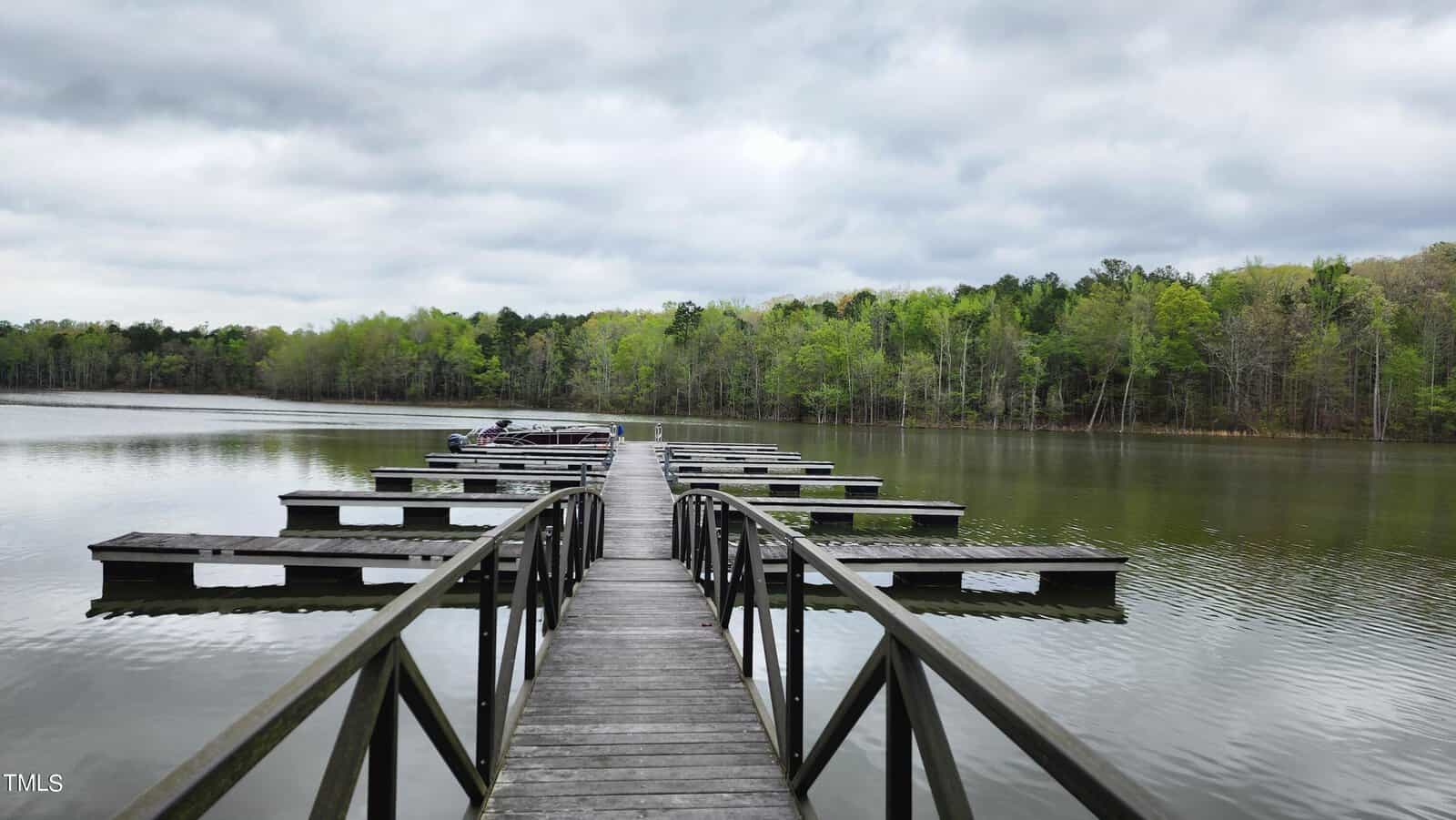 A serene wooden pier extending into a tranquil lake, flanked by lush green trees under an overcast sky. several boats are docked along the pier.