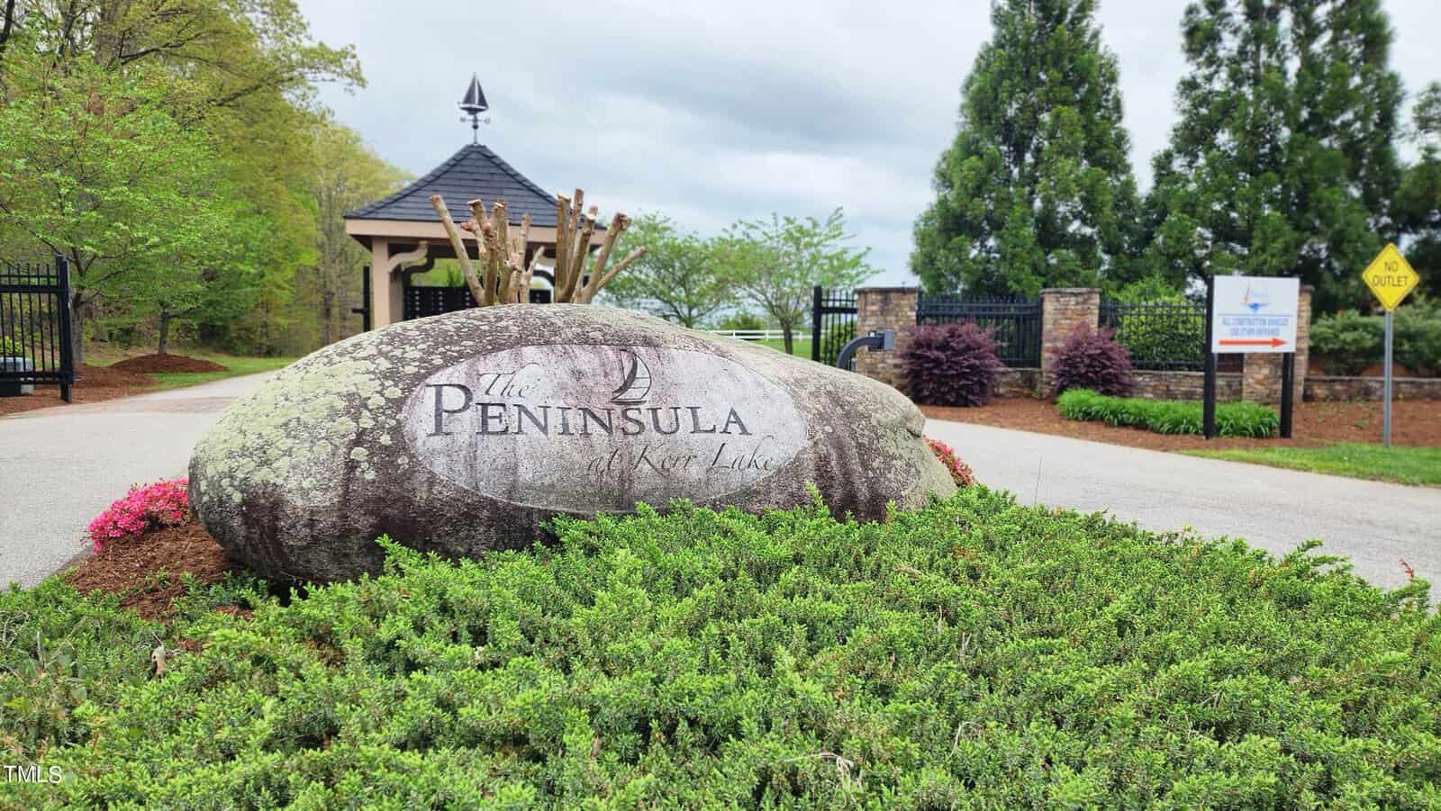 A large engraved rock displaying "the peninsula at kerr lake" sits at the entrance of a gated community, flanked by lush green shrubs and colorful flowers, with a gazebo in the background.