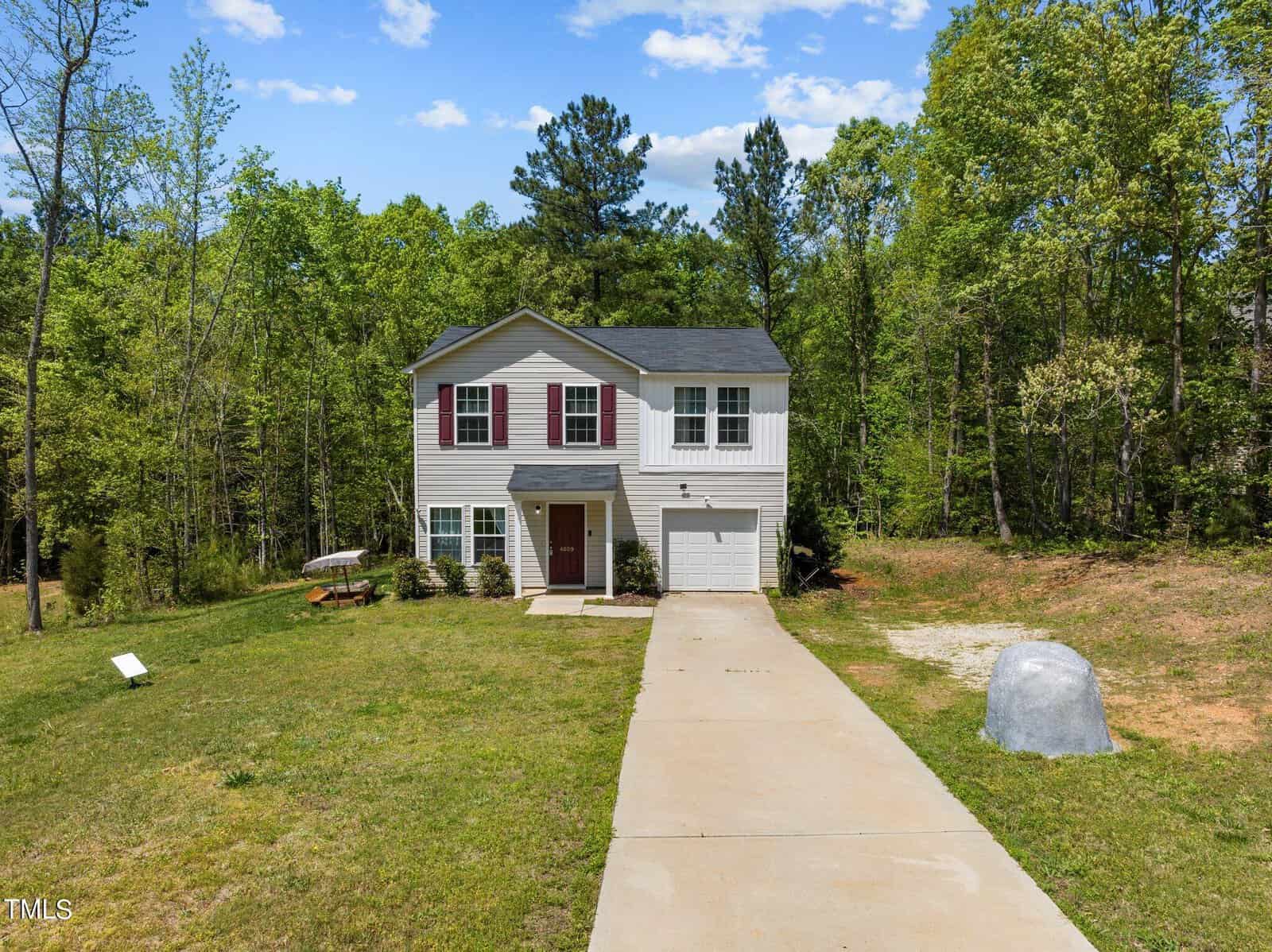 A two-story gray house with red shutters and a neatly maintained lawn. The house has a single car garage and is surrounded by trees and greenery. A long driveway leads up to the front door, and a large boulder is visible on the right side of the driveway.