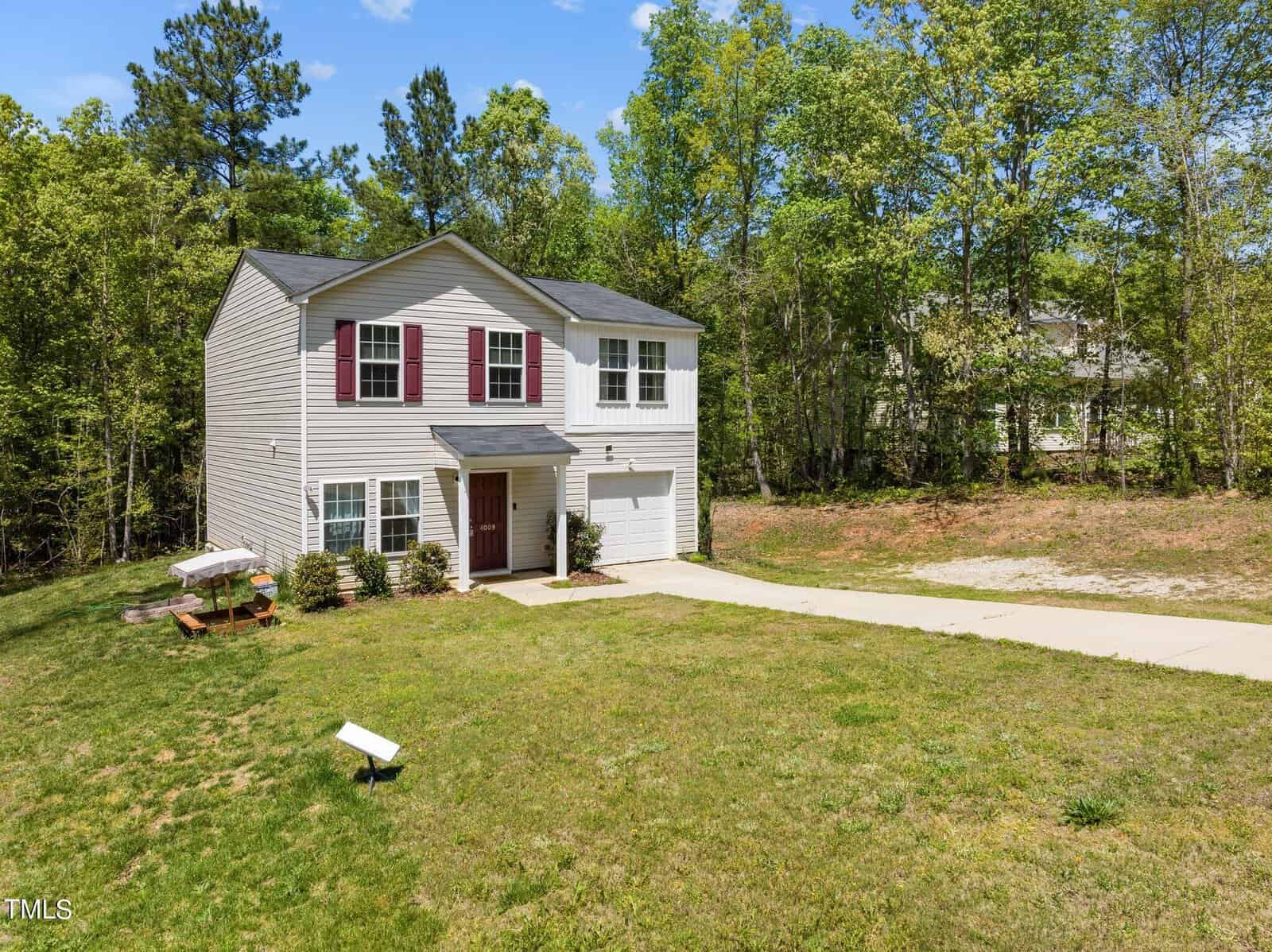 A two-story suburban house with white siding and red shutters, set among green trees. It features a neatly manicured lawn, a single driveway, and a small front porch. There's a picnic table in the front yard and a pathway leading to the main entrance.