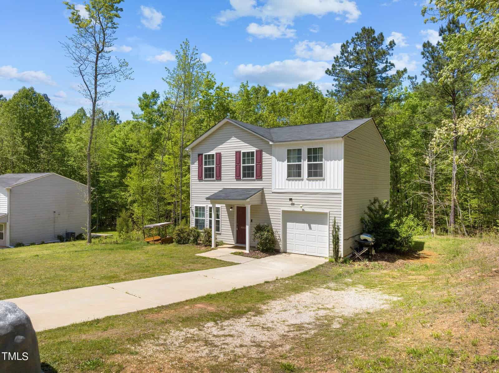 A two-story house with light-colored siding, red shutters, and a single-car garage. It is set amidst a green, wooded area with trees surrounding the property. The house has a concrete driveway and a small, well-kept lawn. The sky is partly cloudy.