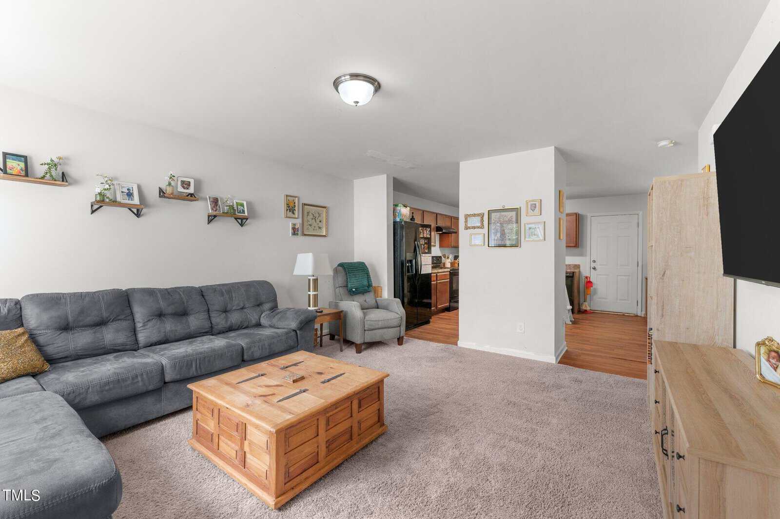 A cozy living room with a large gray sectional sofa, a wooden coffee table, and a TV on a wooden stand. Shelves with framed photos are on the left wall. The room has carpeted flooring and leads into a kitchen with wooden floors in the background.