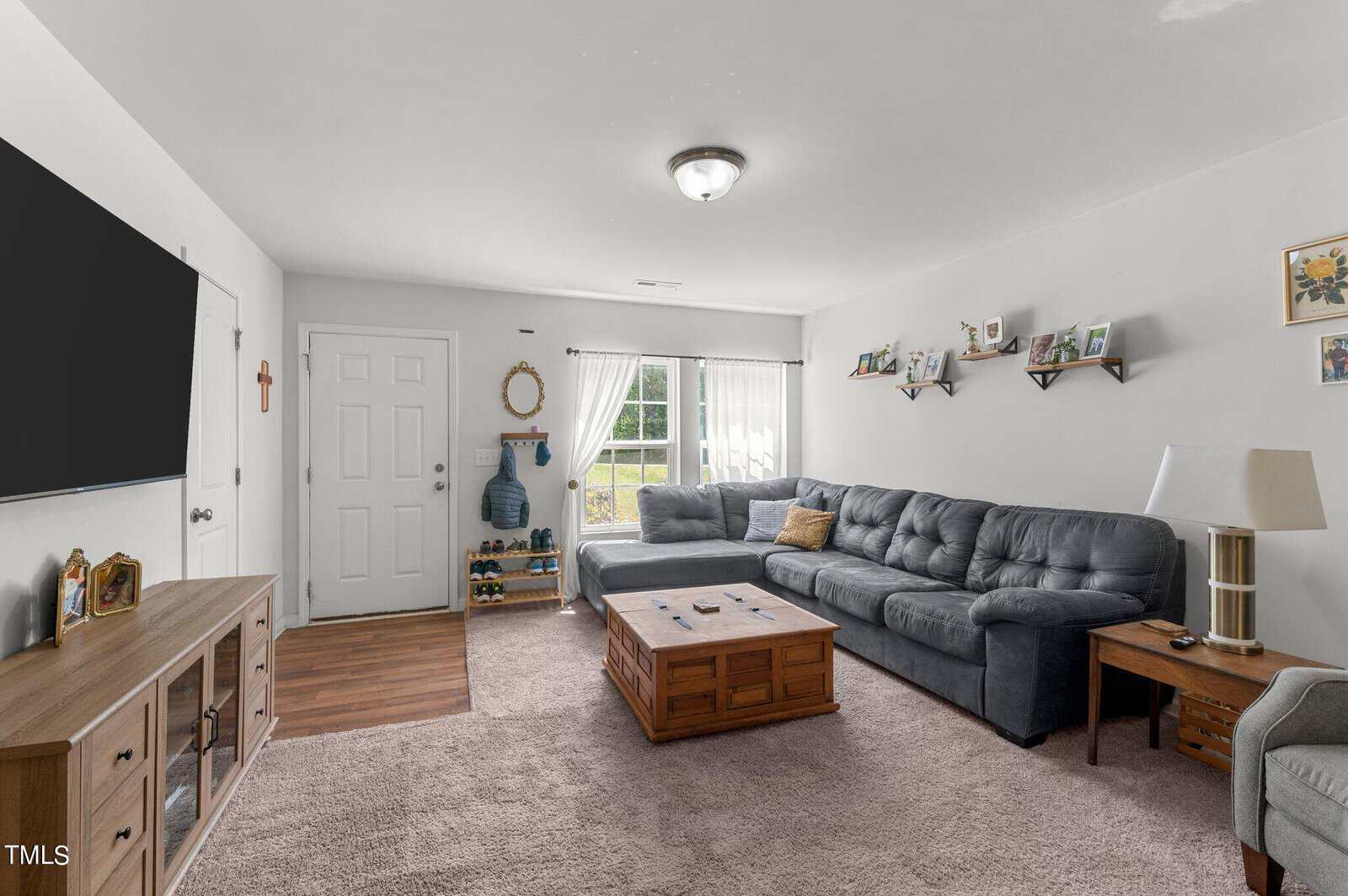 A cozy living room with a large gray sectional sofa, a wooden coffee table, and carpeted floors. The far wall features a TV mounted on a wooden entertainment center. The room is well-lit by natural light from a window with white curtains and a glass door.