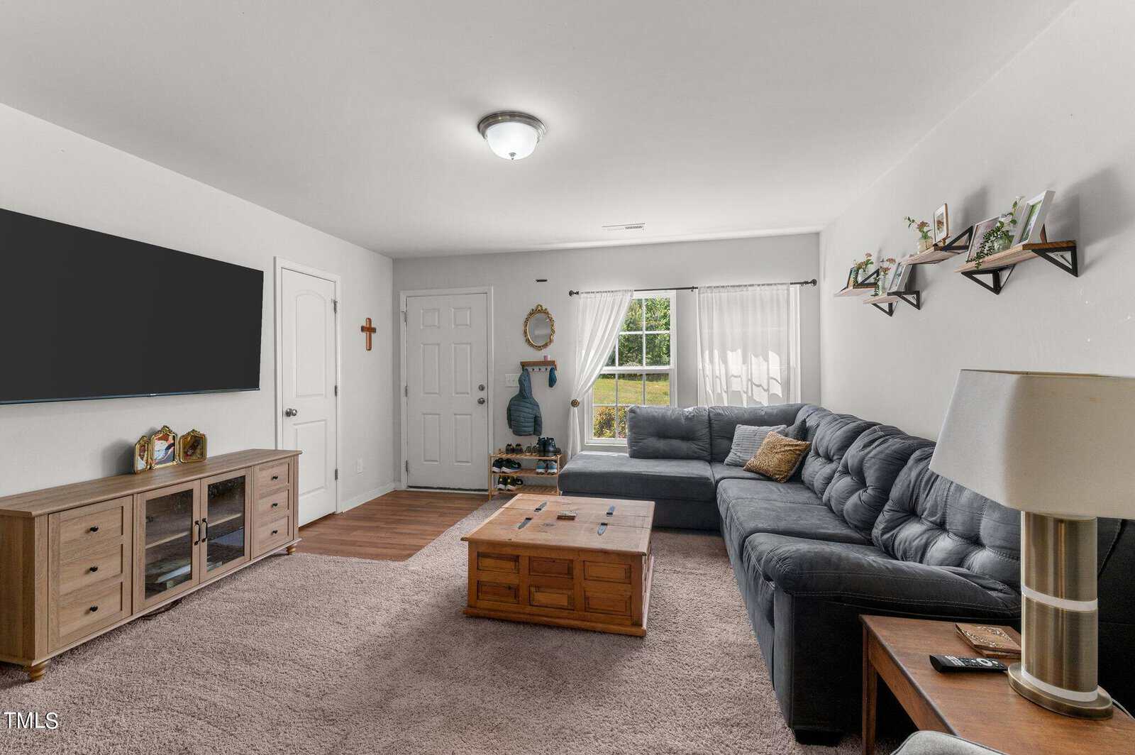 A cozy living room with a gray sectional sofa, a wooden coffee table, and a TV mounted on a white wall. Shelves with decorative items hang above the sofa. A large window with sheer curtains lets in natural light. The wooden floor has a soft beige rug.