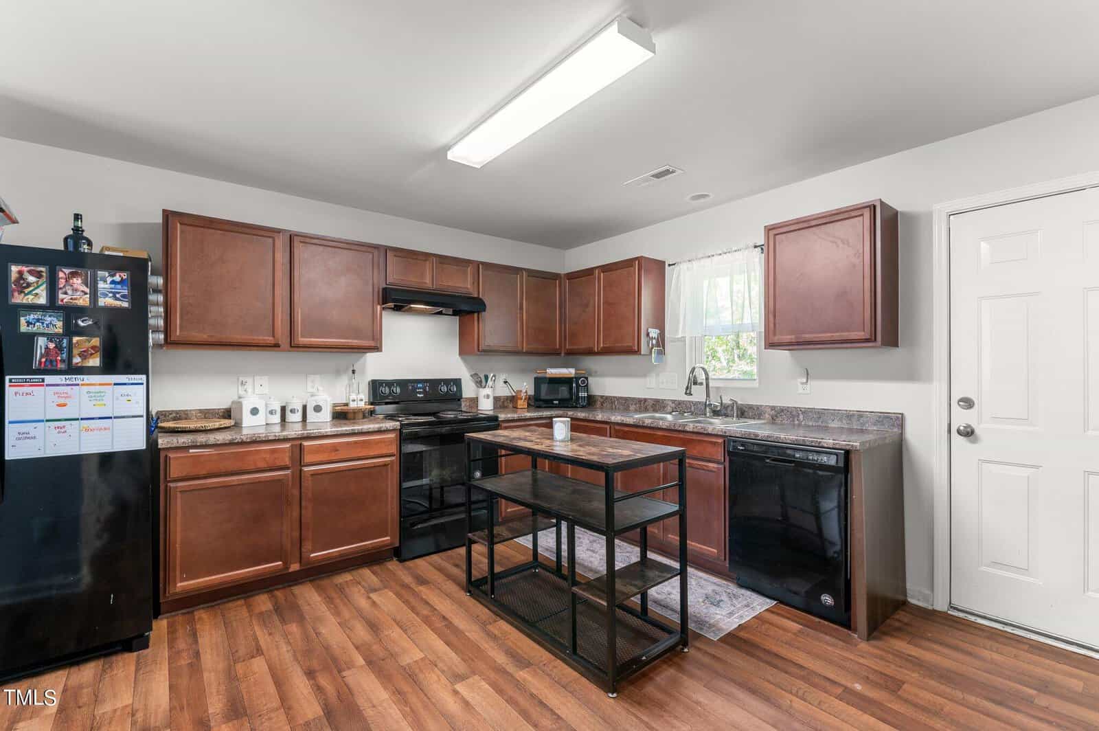 A modern kitchen with wooden cabinets, a black stove, black dishwasher, and a black fridge adorned with magnets. A small island with a metal frame and glass shelves sits in the center. The floor is wooden, and the walls are white. A window with a white curtain provides natural light.