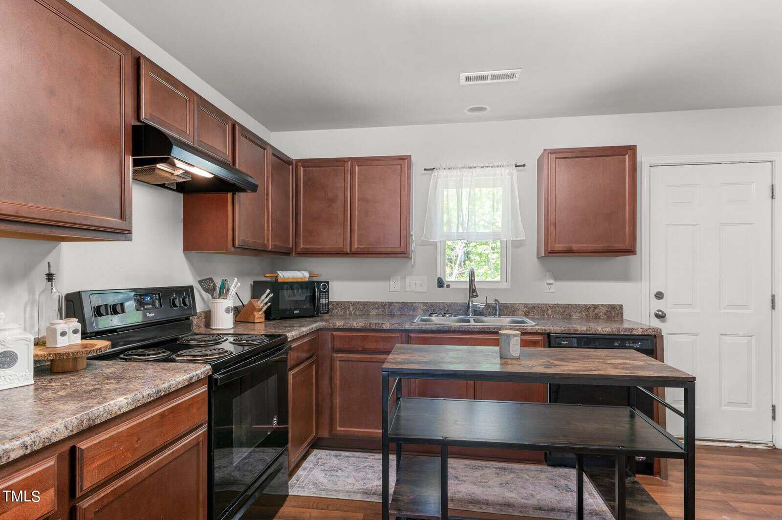 A modern kitchen featuring wooden cabinets, a marbled countertop, and black appliances, including a stove, oven, and dishwasher. A window with sheer curtains is above the sink, and a central island with a black metal frame and dark wooden top is in the middle.