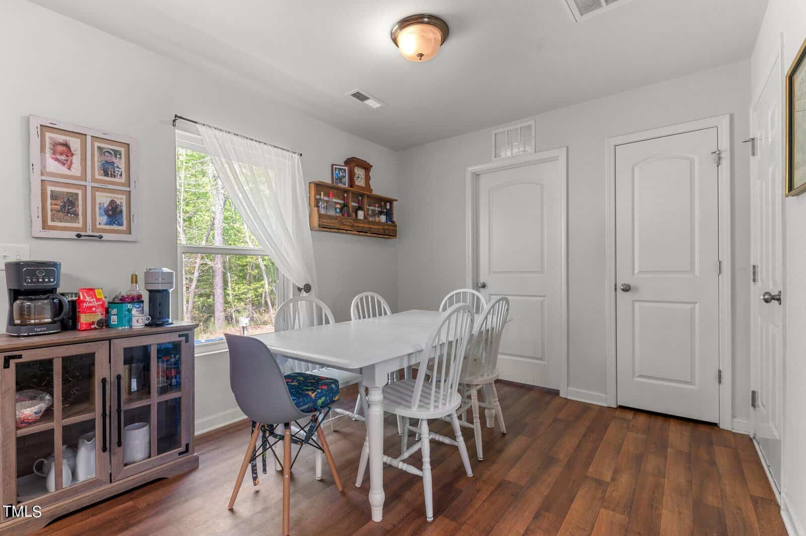 A bright dining room with a wooden floor features a white rectangular table surrounded by white chairs and one gray chair. A small sideboard with a coffee maker and beverages is on the left, and two closed white doors are on the right wall. A window with sheer white curtains is in the background, next to wall-mounted picture frames.