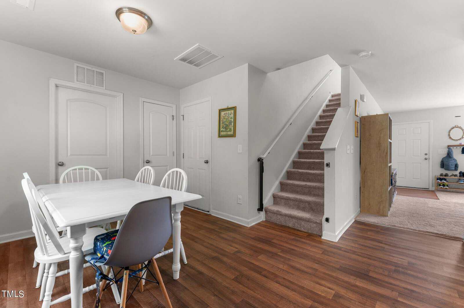 A room with wooden flooring featuring a white dining table with four chairs, one being grey. Adjacent is a carpeted staircase leading upward. The background shows closed white doors and an open space leading to another room, with a cabinet visible.