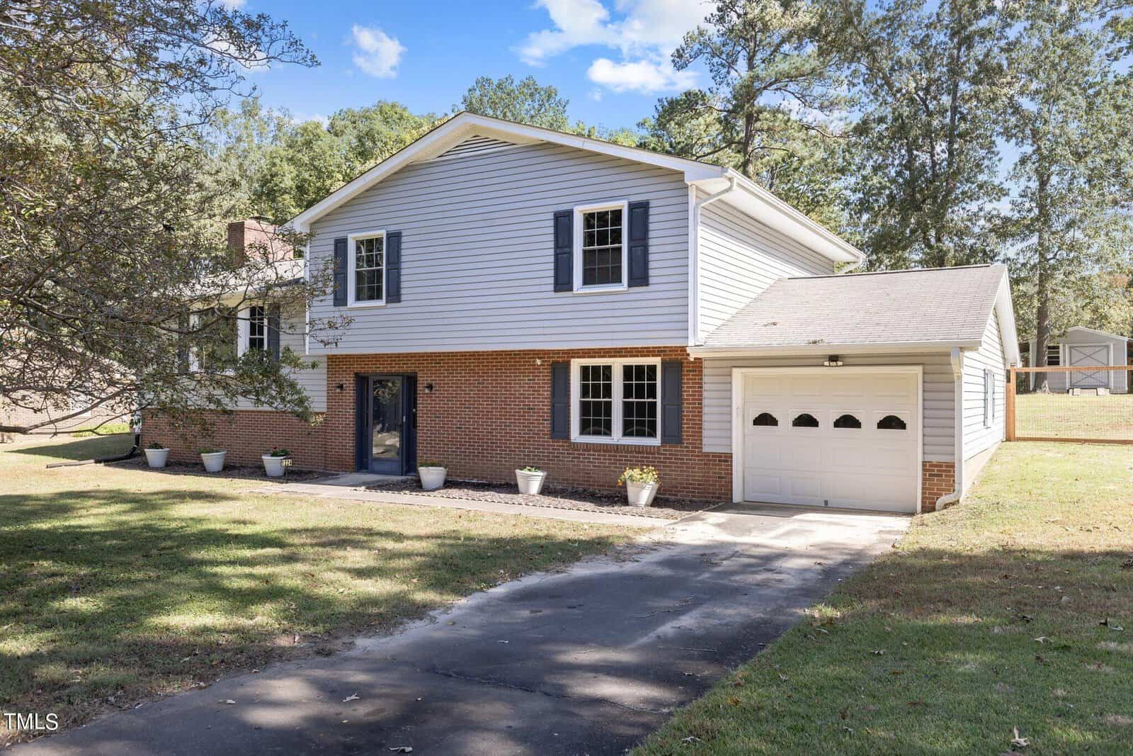 The charming two-story house at 1224 Hedrick Drive in Henderson boasts red brick and white siding, set in a grassy yard. It features a single-car garage, blue shutters, and several potted plants lining the walkway to the front door, with trees gracing the background under a clear blue sky.