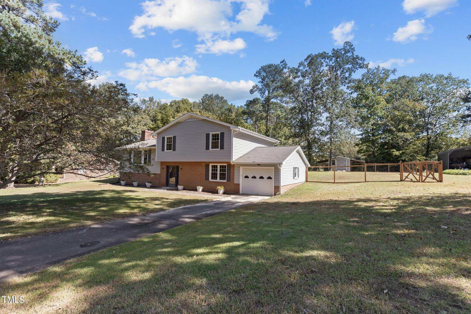 A two-story suburban house with a brick and siding exterior, two-car garage, and a front yard with trees. The house has a driveway and a fenced backyard, surrounded by greenery under a partly cloudy sky.