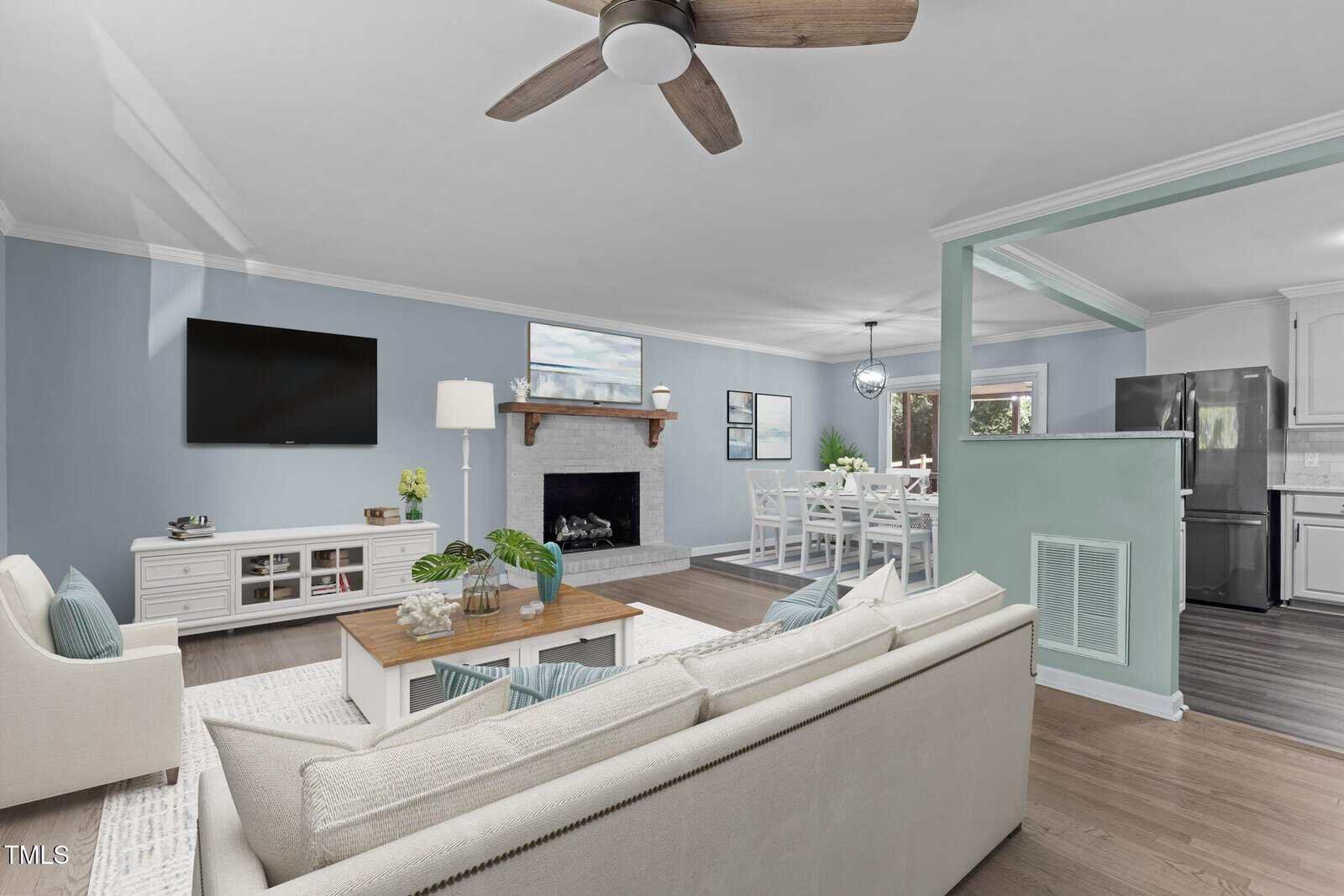 A modern living room with a beige sofa, wooden coffee table, and a fireplace. The room features light blue walls, a ceiling fan, and a wall-mounted TV. An open dining area is visible in the background, with a view into the kitchen.