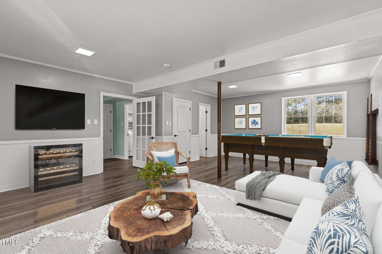A spacious, modern living room with a large TV on the wall. A pool table is near the windows, and there's a wooden coffee table surrounded by a white sectional sofa. The room has grey walls, hardwood flooring, and a patterned rug.